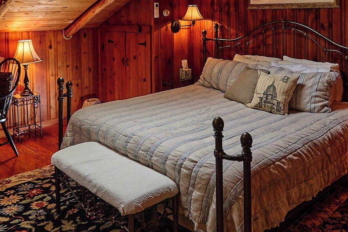 A cozy attic bedroom with a wooden ceiling, a skylight, a bed with a bench, and a lamp.