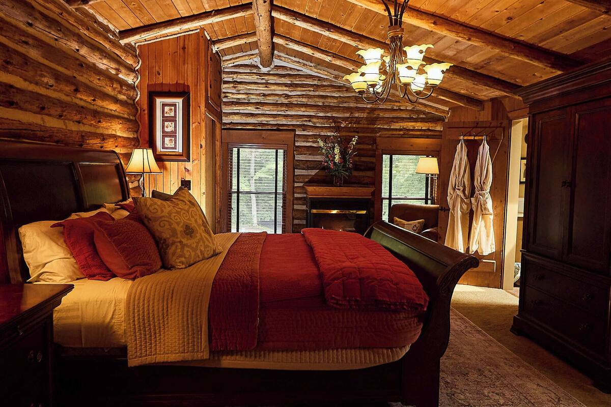 A log cabin bedroom with a large bed, wooden walls, a chandelier, and a fireplace.