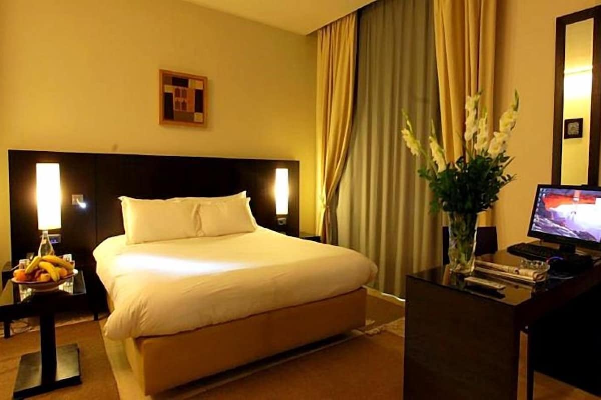 A hotel room with a large bed, a desk with a computer, a vase with flowers, and a window with curtains.