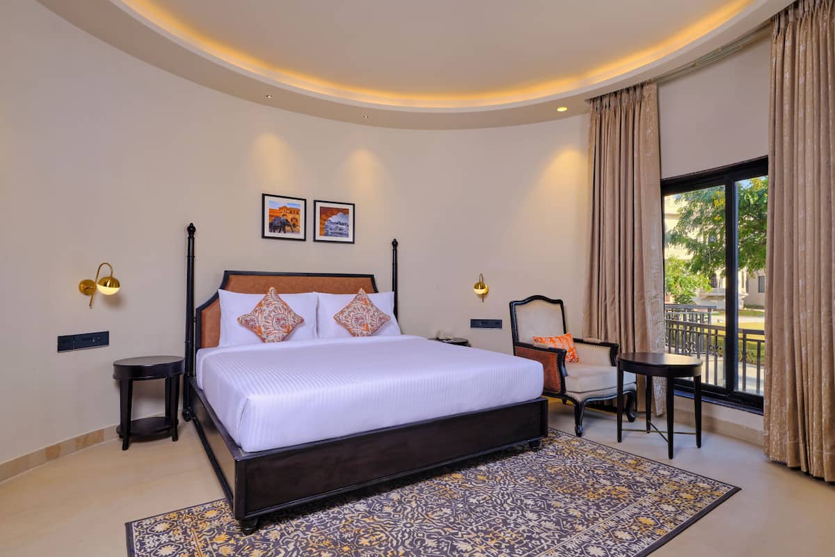 Quarto de hotel com uma cama grande, duas luzes fixadas na parede, uma mesa pequena, uma cadeira e uma varanda com cortinas.