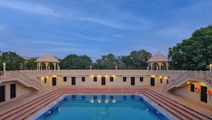 Una grande piscina rettangolare con gradini che conducono a una zona coperta, circondata da un edificio bianco con numerose finestre e una ringhiera decorativa.