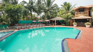 A swimming pool with lounge chairs and umbrellas, surrounded by palm trees and a building with a balcony. - Ruffles Beach Resort (Candolim)