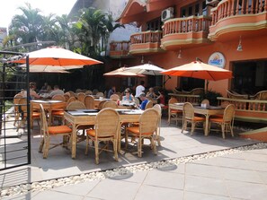 An outdoor dining area with wicker tables and orange cushions, surrounded by a low stone wall and palm trees. - Ruffles Beach Resort (Candolim)