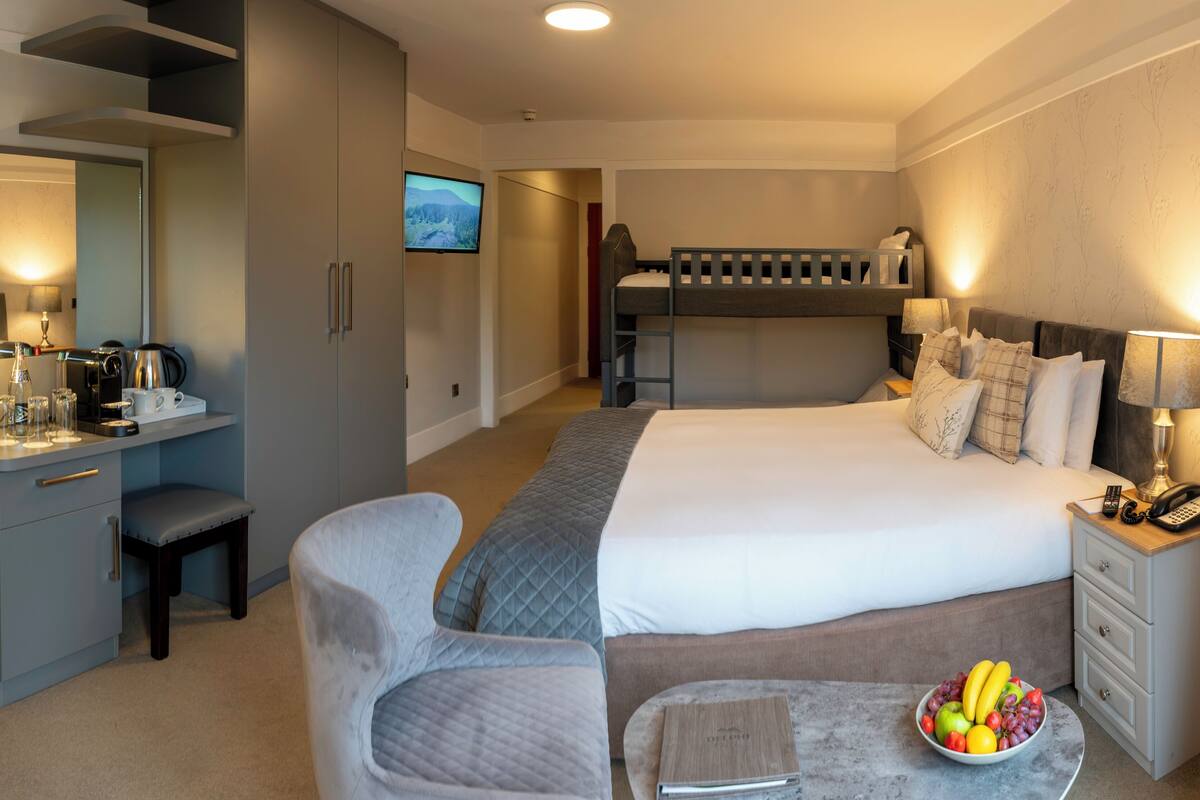 A hotel room with a bed, desk, chair, and a small table with a fruit bowl.