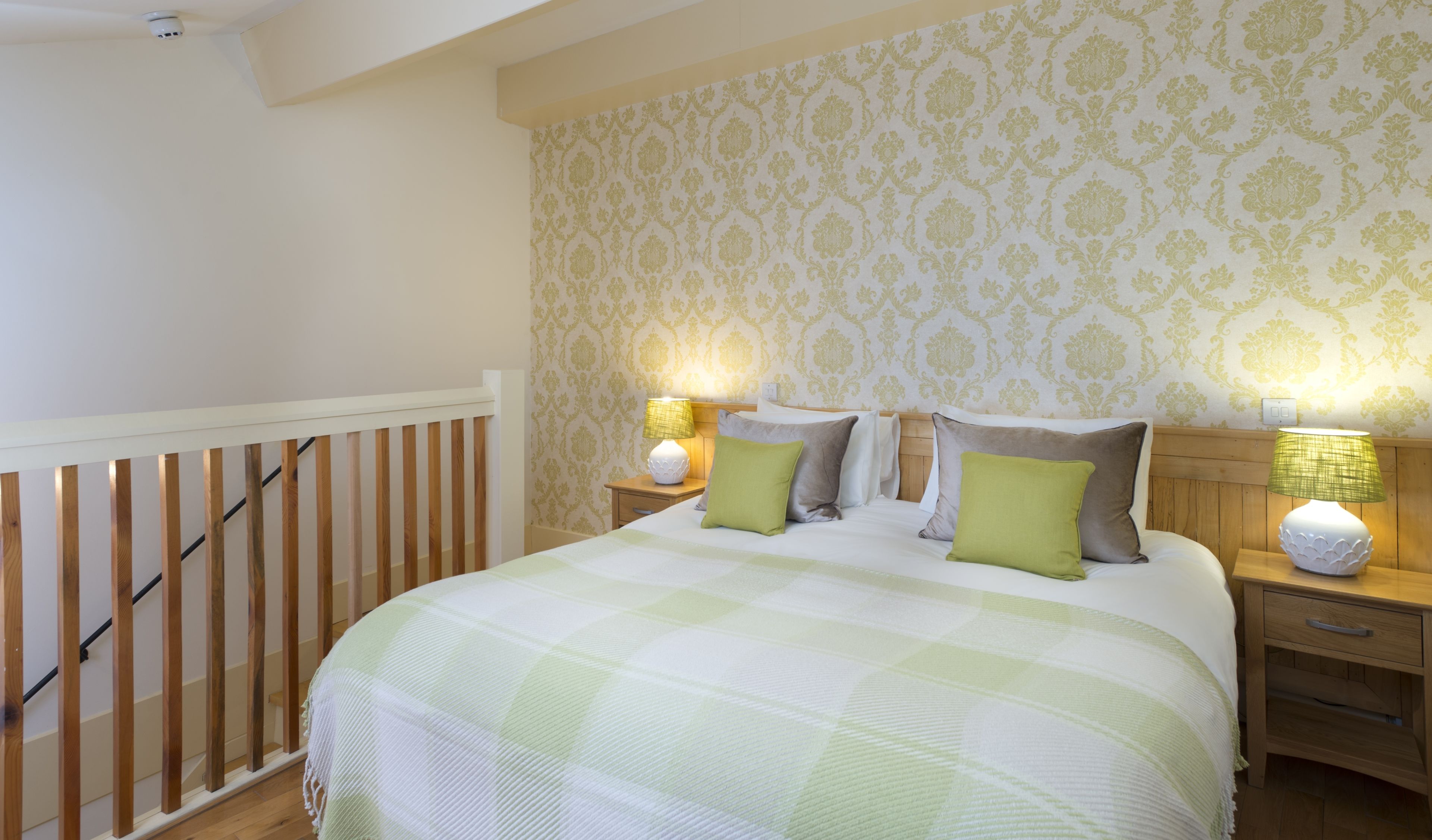 A bedroom with a bed, two bedside tables, a nightstand, a wooden railing, and patterned wallpaper.