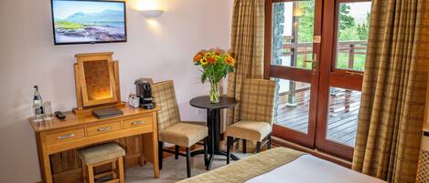 A hotel room with a bed, a desk with a computer, two chairs, a small table with a vase of flowers, a television on the wall, and a large window with curtains.