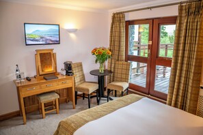 A hotel room with a bed, a desk with a computer, two chairs, a small table with a vase of flowers, a television on the wall, and a large window with curtains.