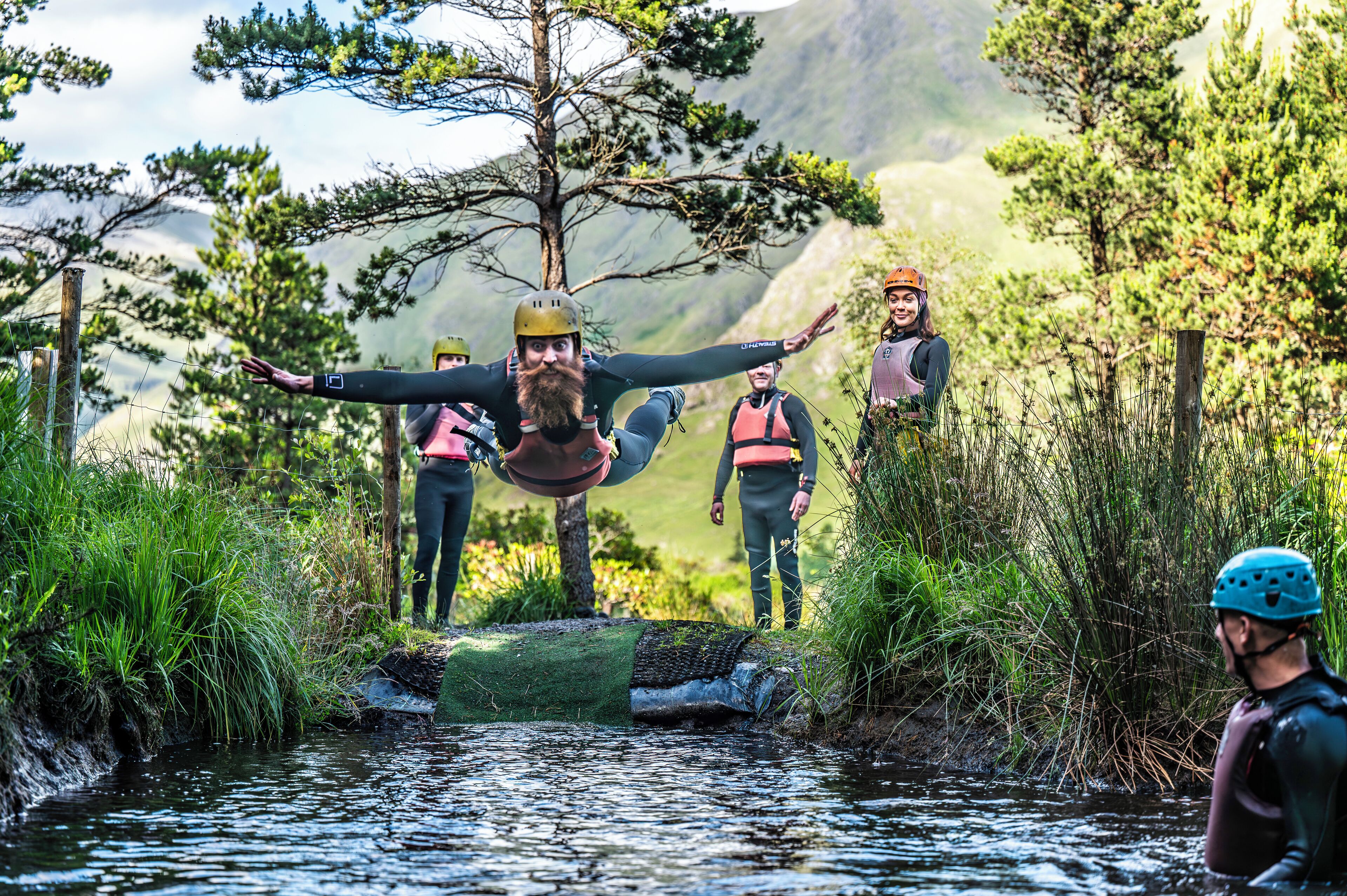 Un groupe de personnes, vêtues d’habits adaptés aux activités de plein air, se trouve près d’une rivière, avec des arbres et des montagnes en arrière-plan.