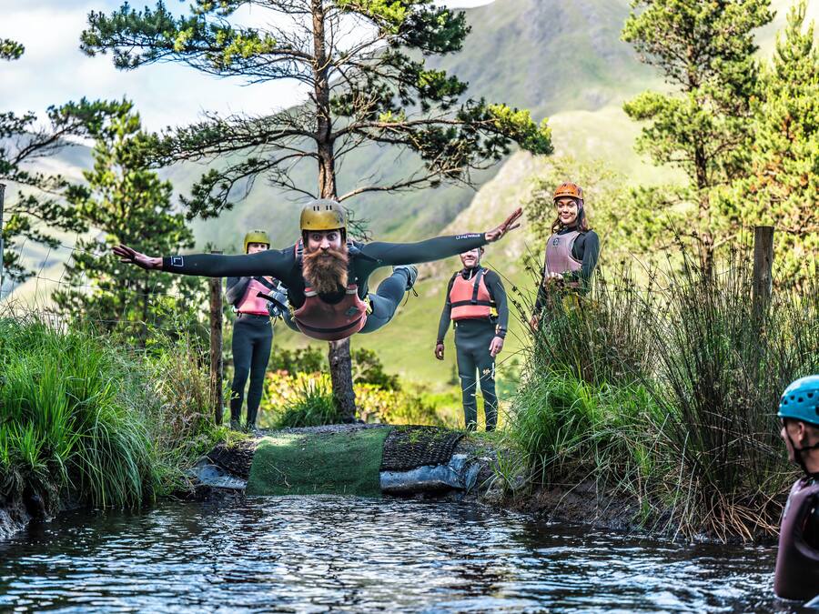 Un grupo de personas con equipo de senderismo junto a un río, con árboles y montañas al fondo.