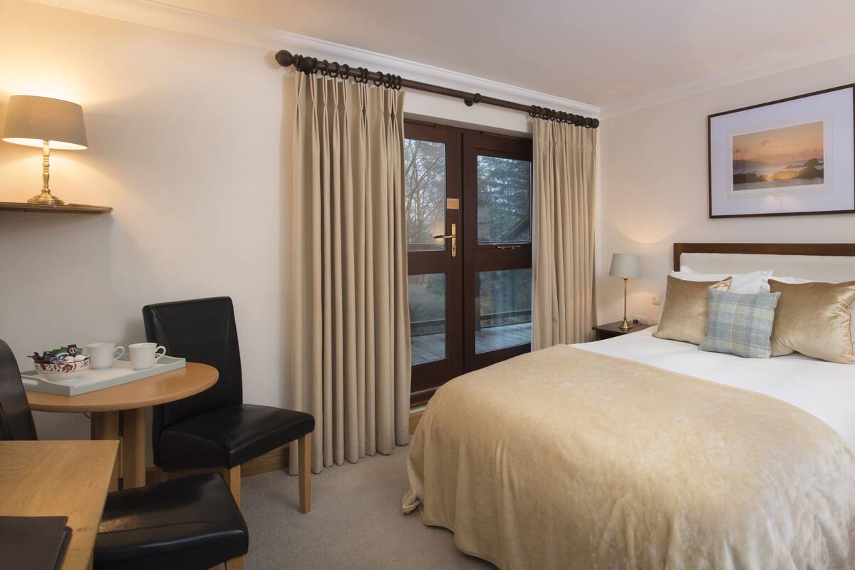 A hotel room with a bed, a desk, a chair, a window with curtains, a bedside table with a lamp, and a framed picture on the wall.