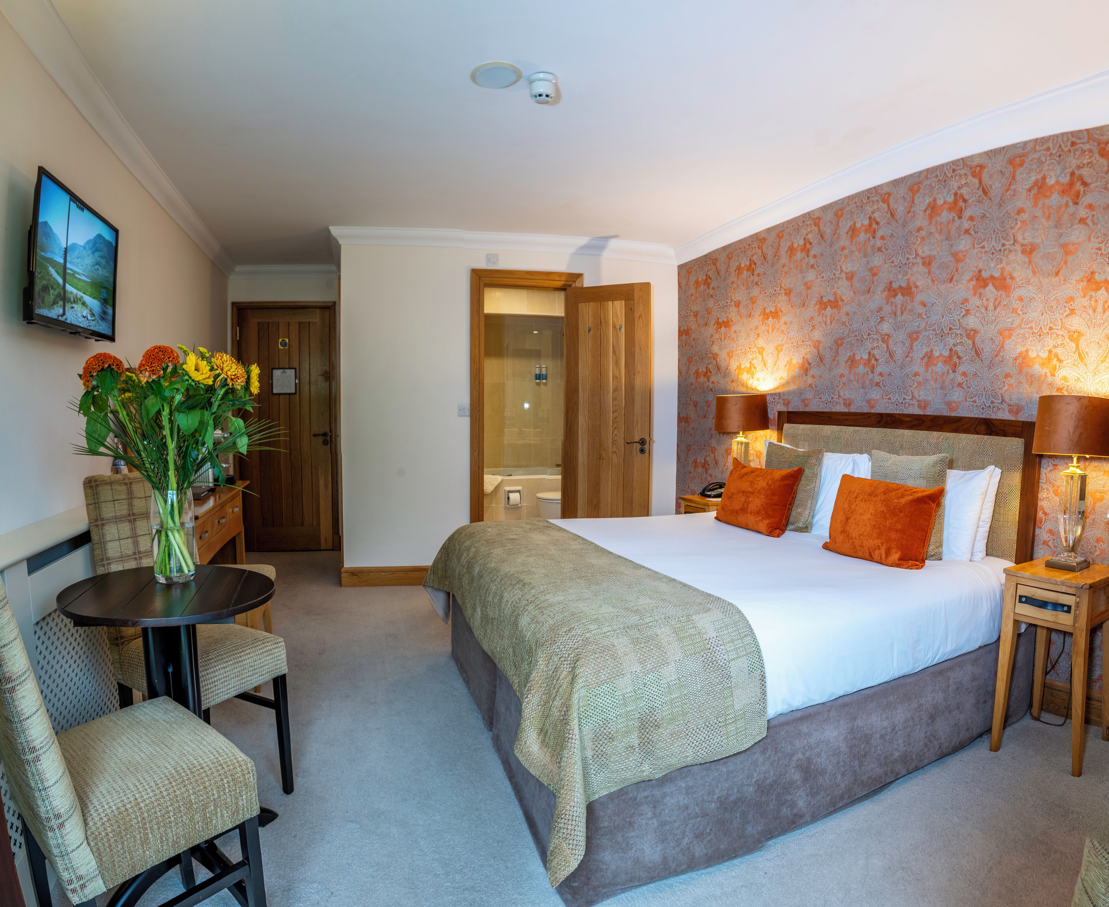 A hotel room with a large bed, two bedside tables with lamps, a chair, a small table with a vase of flowers, and a bathroom visible through an open door.