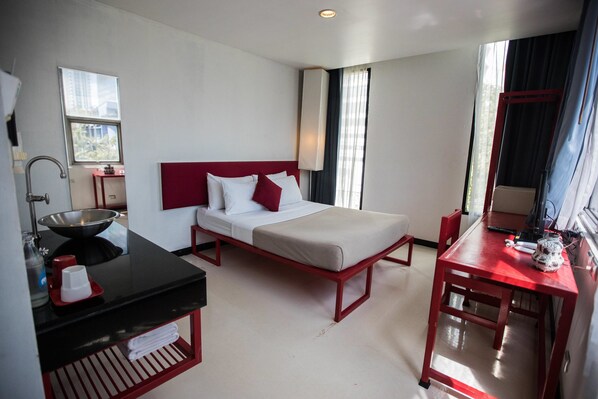 A hotel room with a bed, a desk, a red-framed mirror, a sink, and a window with curtains.
