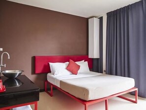 A hotel room with a red bed frame, white bedding, a small bedside table with a red lamp, a black desk with a silver bowl, and a window with dark curtains.