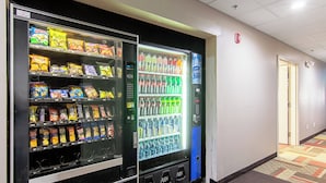 A vending machine with snacks and drinks, located in a hallway with a carpeted floor and a door in the background. - Red Roof Inn Locust Grove (Locust Grove)