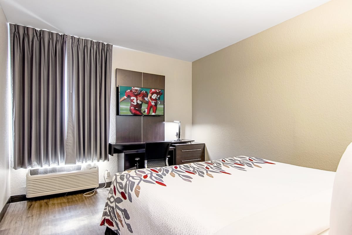 A hotel room with a large bed, a flat-screen TV mounted on the wall, a desk with a chair, and a window with curtains.