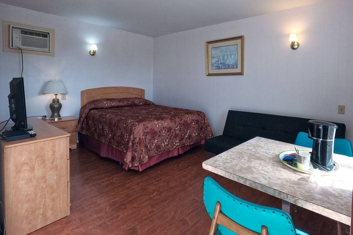 A room with a bed, a desk with a lamp, a table with a chair, a painting on the wall, and a coffee machine.