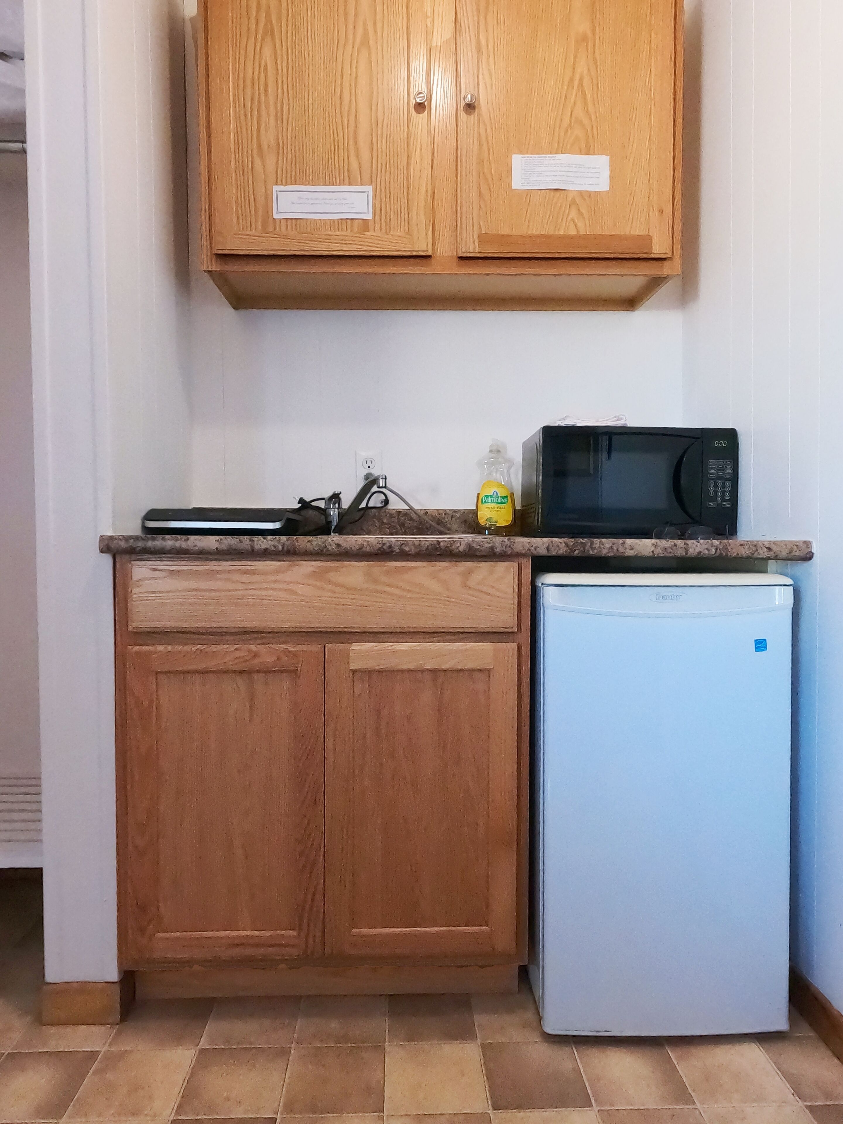 A kitchen with wooden cabinets, a microwave, a small refrigerator, and a bottle of dish soap on the countertop.