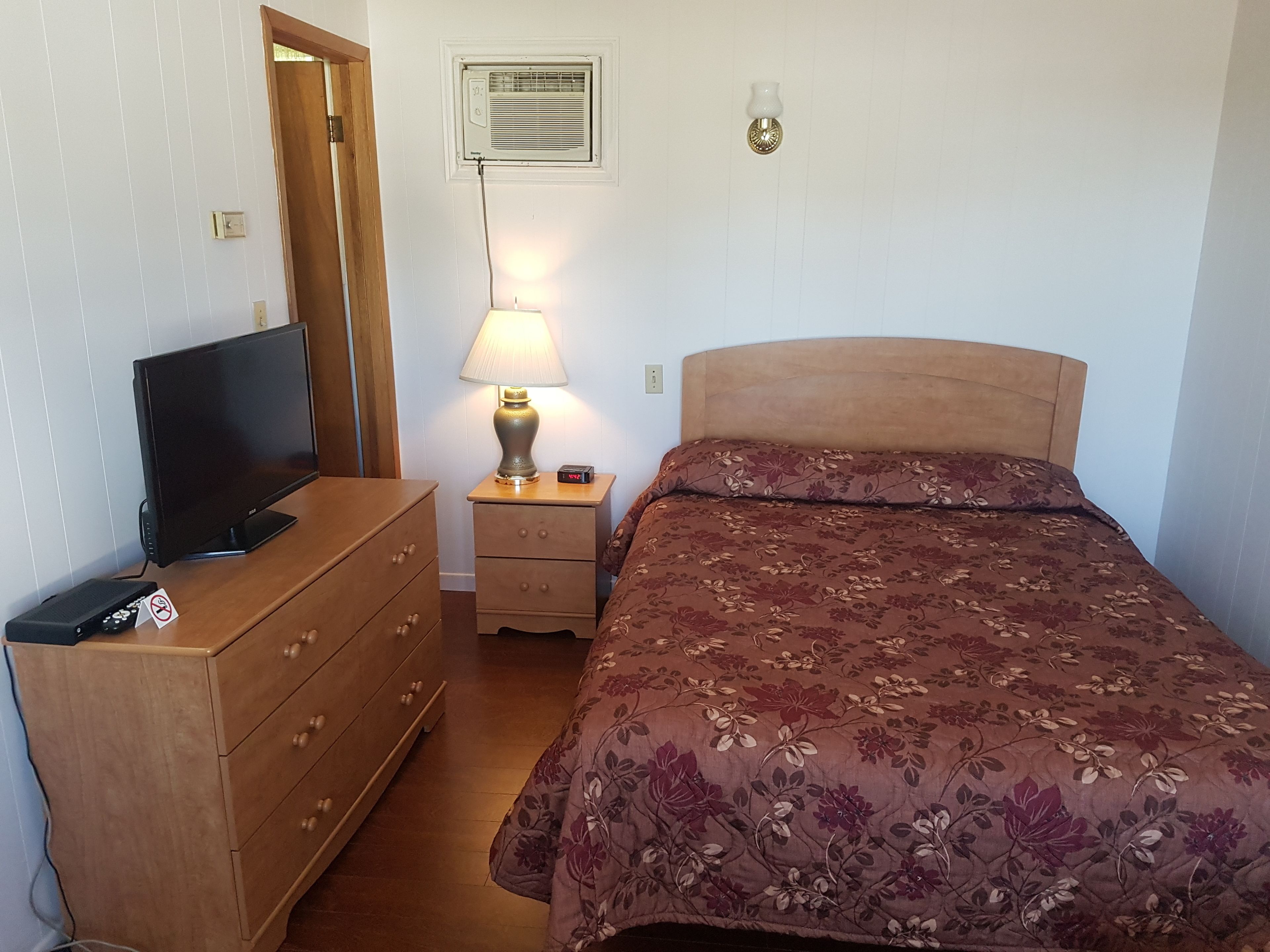 A bedroom with a bed, a dresser, a TV, a lamp, and a wall-mounted air conditioner.