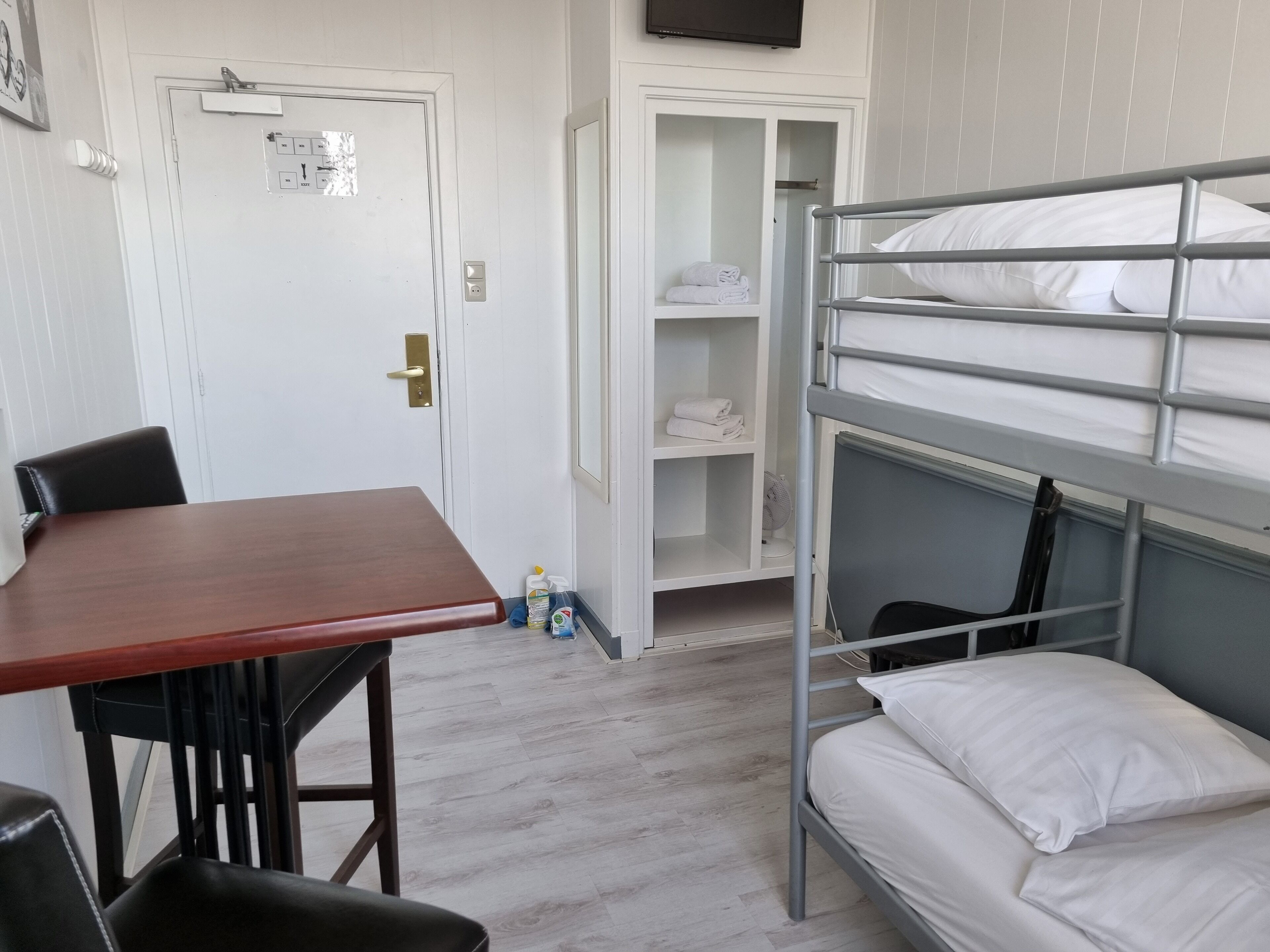 A bunk bed with white bedding, a wooden table, and a chair in a room with white walls and a tiled floor.