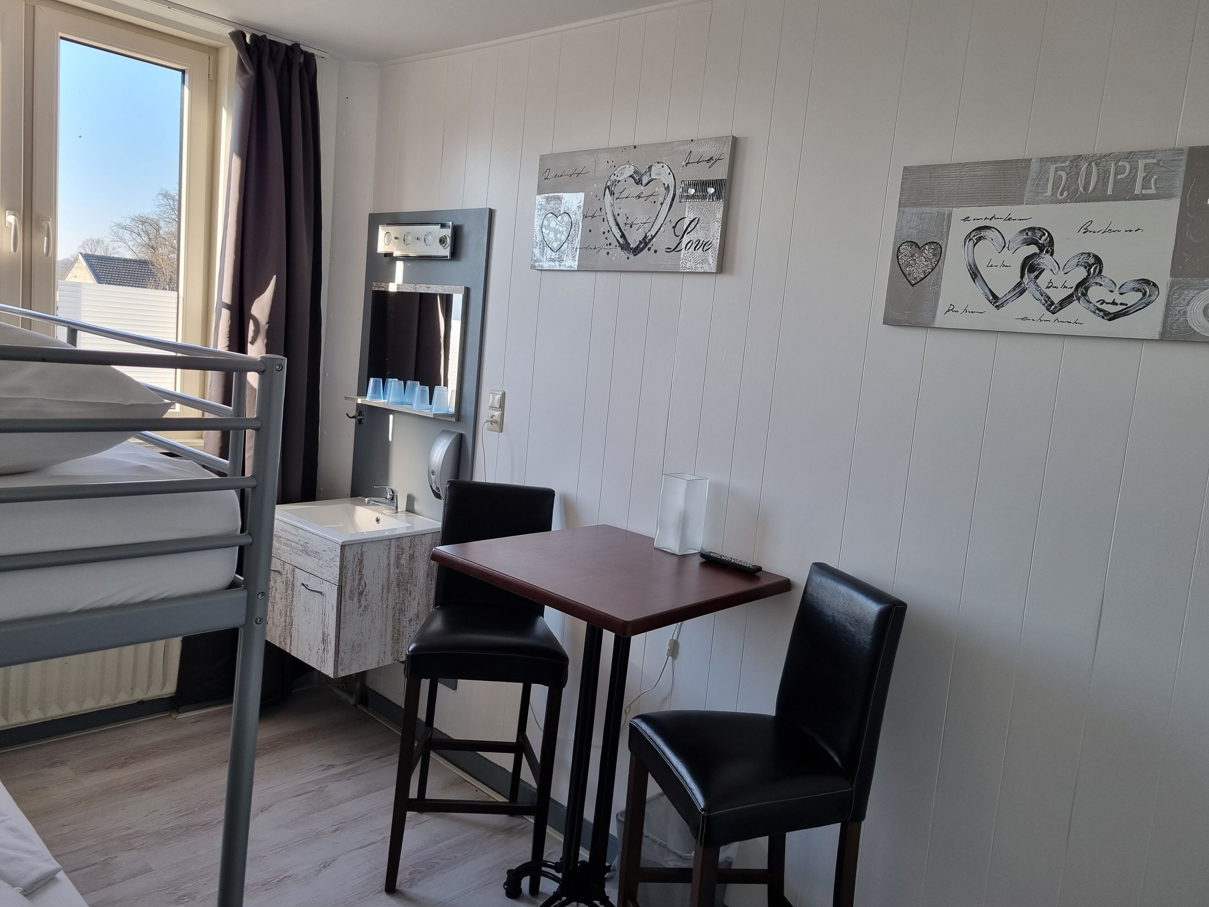 A room with bunk beds, a desk, a chair, and a small table. There are wall decorations and a window with curtains.