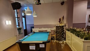 A pool table in a room with wooden flooring, a white cushioned bench, and a decorative plant arrangement.