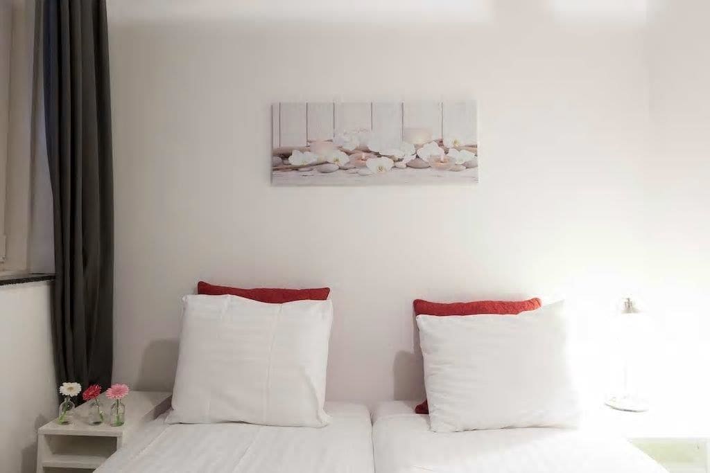 Habitación de hotel con dos camas, ropa de cama blanca, almohadas rojas, una mesita de noche con un florero y un cuadro en la pared.