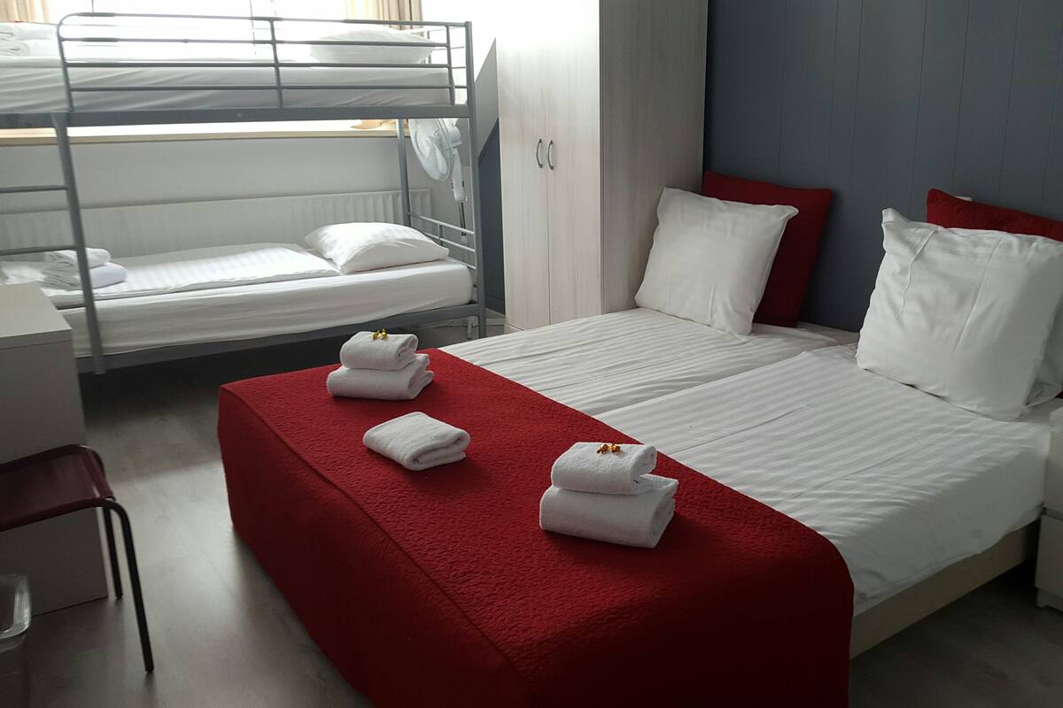 Une chambre d’hôtel avec un lit superposé, un lit simple et un bureau. Il y a des serviettes sur le lit et une lampe sur le bureau.