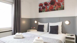 A hotel room with a large bed, two bedside tables, a painting of red flowers, and a window with curtains.