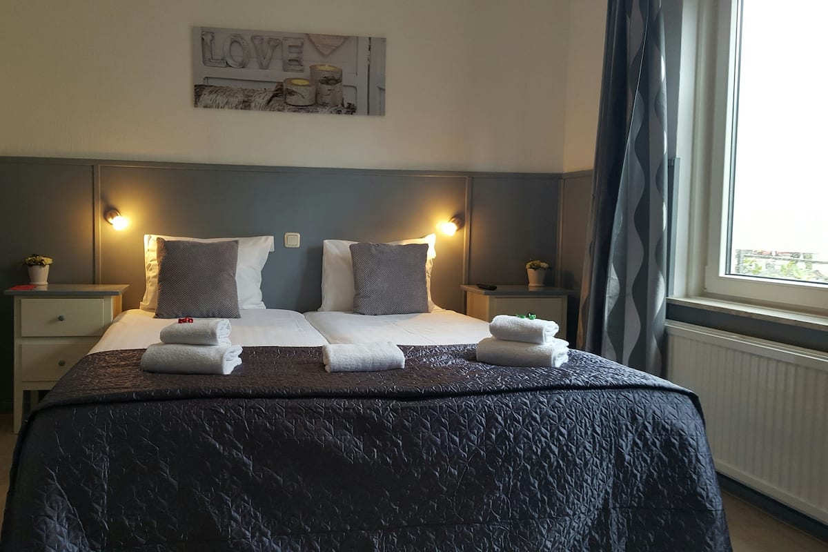 Un lit bien fait, avec deux oreillers, une couverture sombre et deux serviettes pliées. Une lampe est fixée au mur au-dessus du lit, et une petite table avec une plante se trouve à côté.