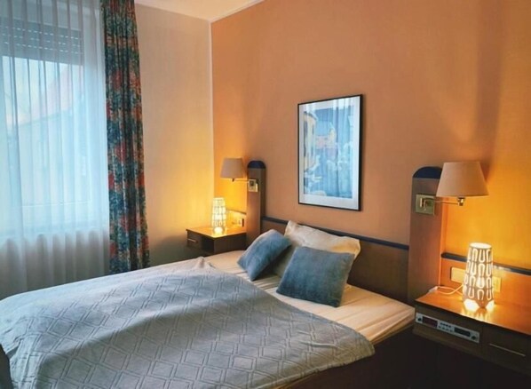 A hotel room with a bed, two bedside lamps, a framed picture on the wall, and a window with curtains. - Hotel Krasemann (Isselburg)