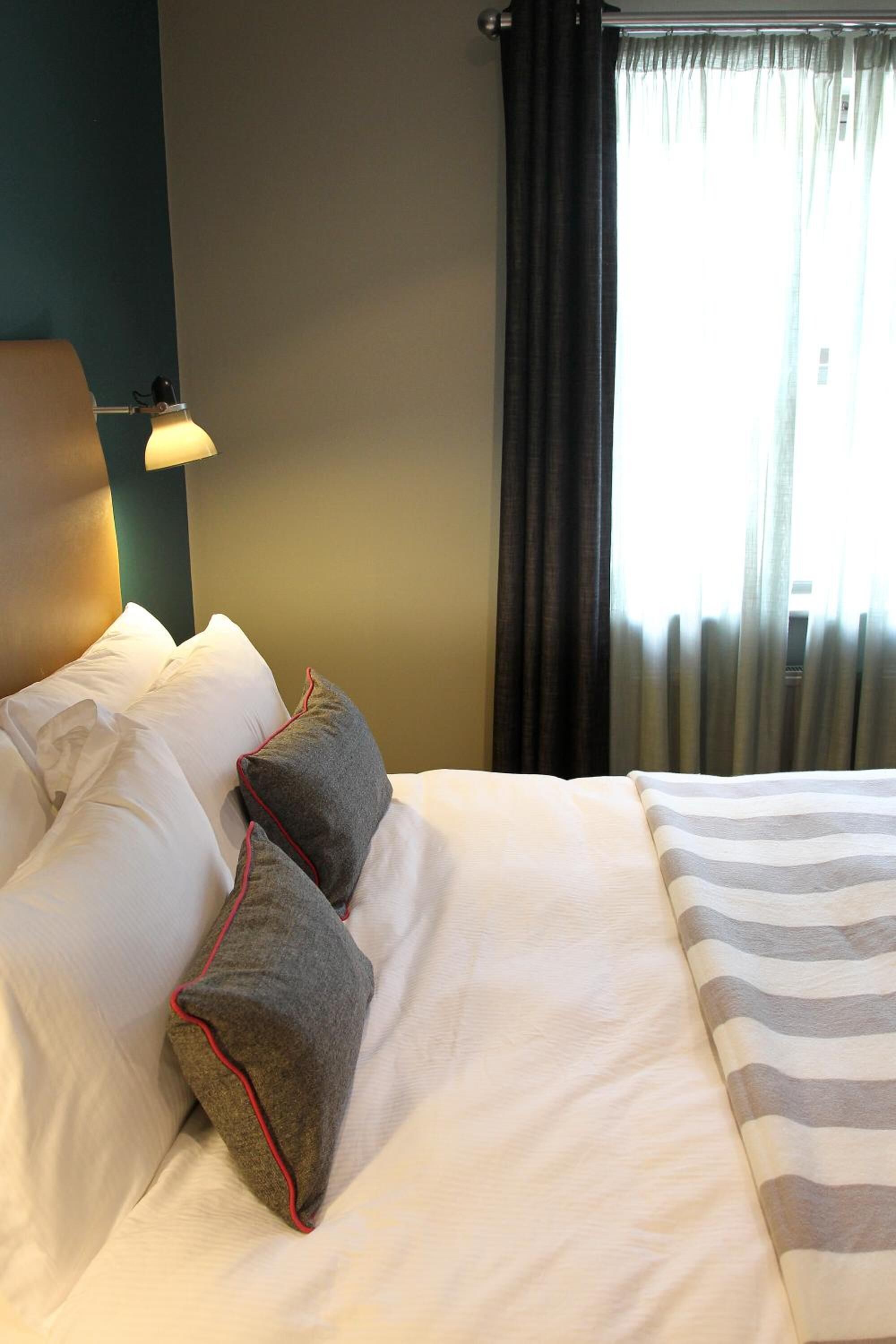 Una cama bien hecha con ropa de cama blanca y a rayas, una lámpara a la izquierda y una ventana con cortinas.