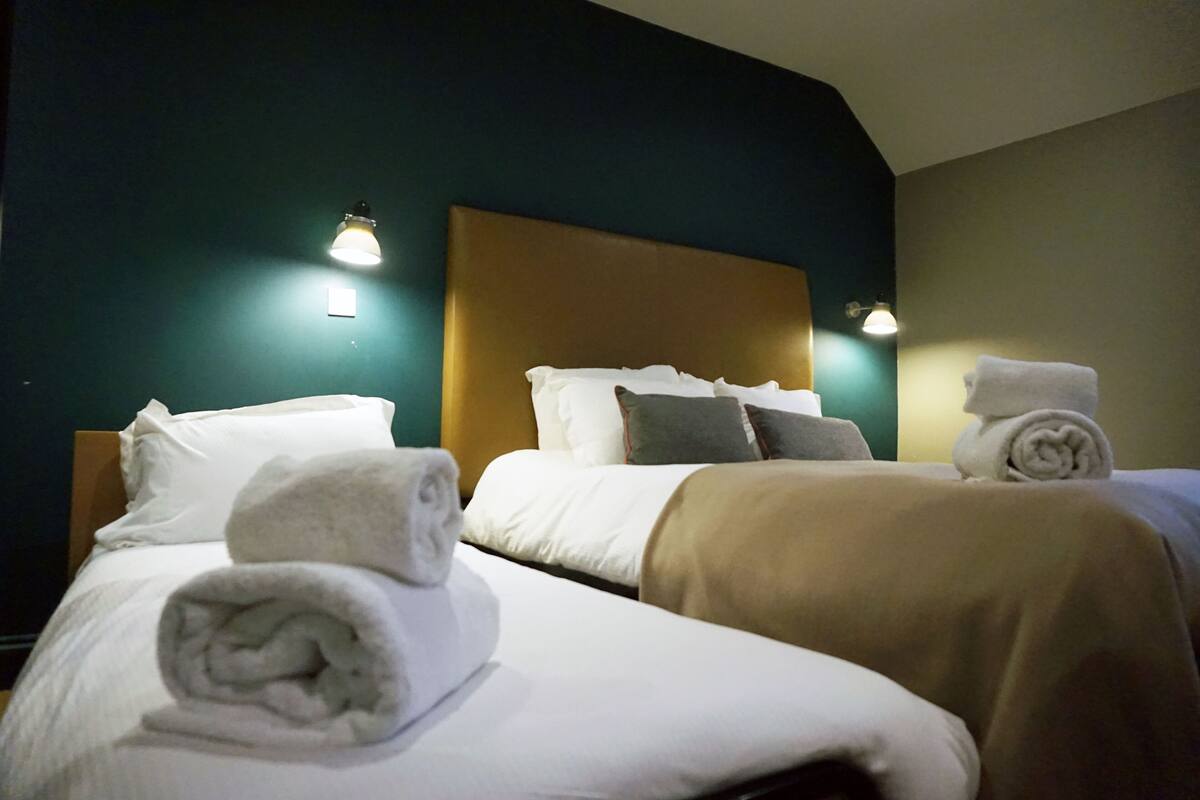 A hotel room with two beds, each with a rolled towel and a pillow. There are wall-mounted lights on either side of the beds.