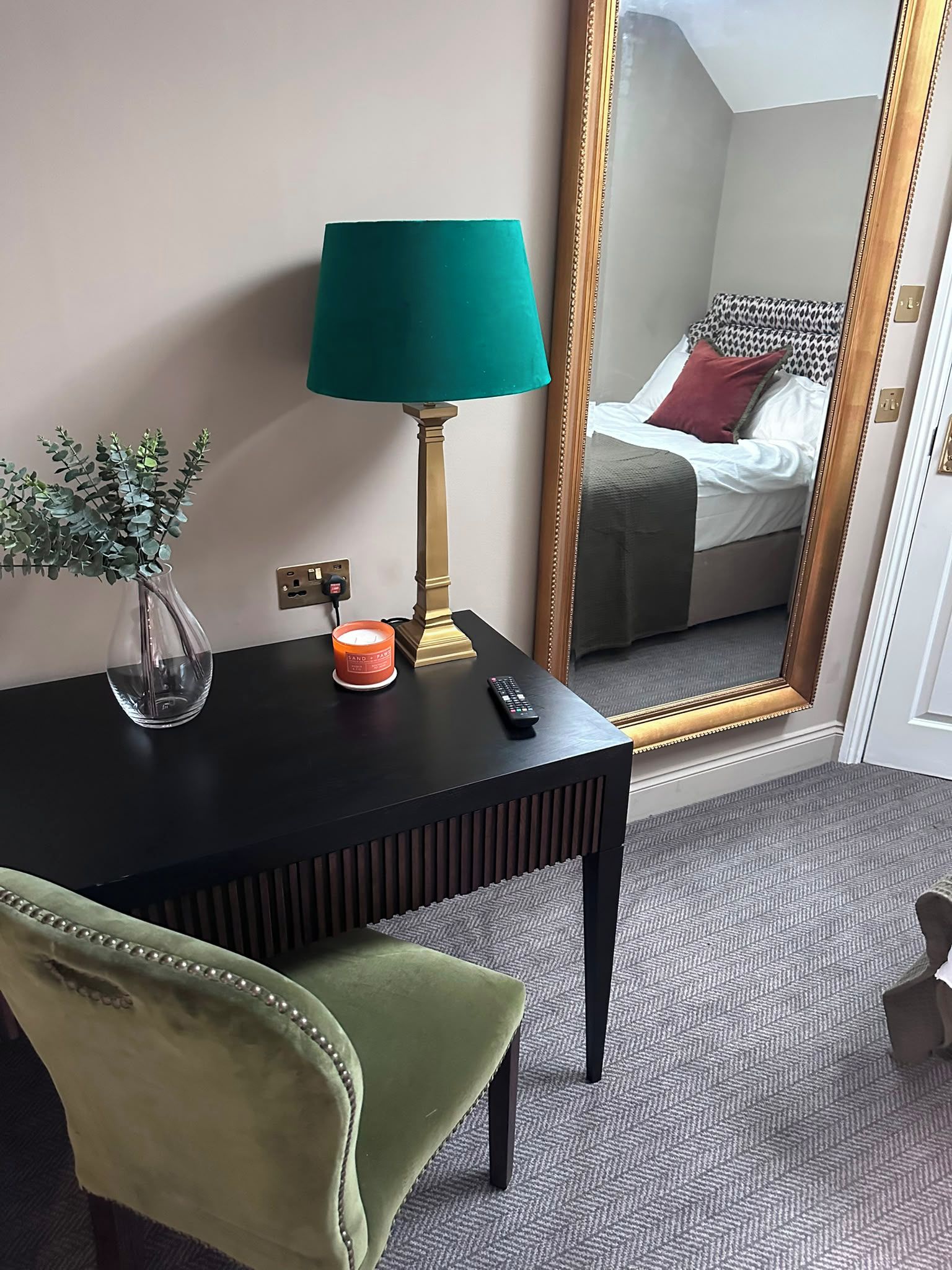 A hotel room with a bed, a mirror, a desk with a lamp, a vase with a plant, and a remote control.