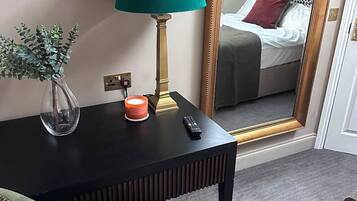 A hotel room with a bed, a mirror, a desk with a lamp, a vase with a plant, and a remote control.