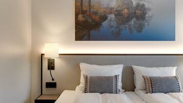 Una cama bien hecha con sábanas blancas y almohadones estampados, una mesita de noche con una lámpara, y un cuadro grande con un paisaje boscoso en la pared.