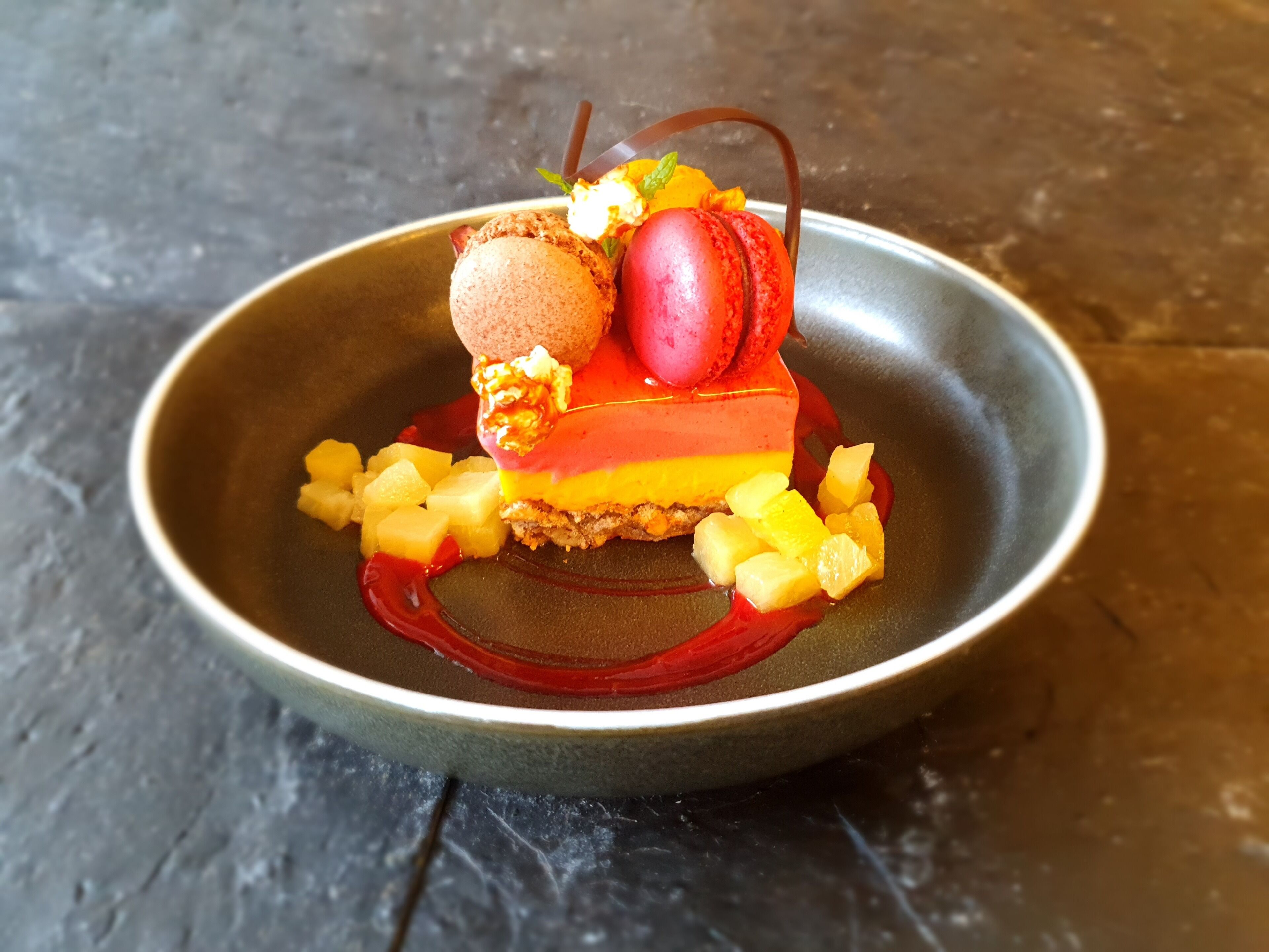 A dessert plate with a layered cake, a scoop of ice cream, a macaron, and diced fruit on a dark plate.