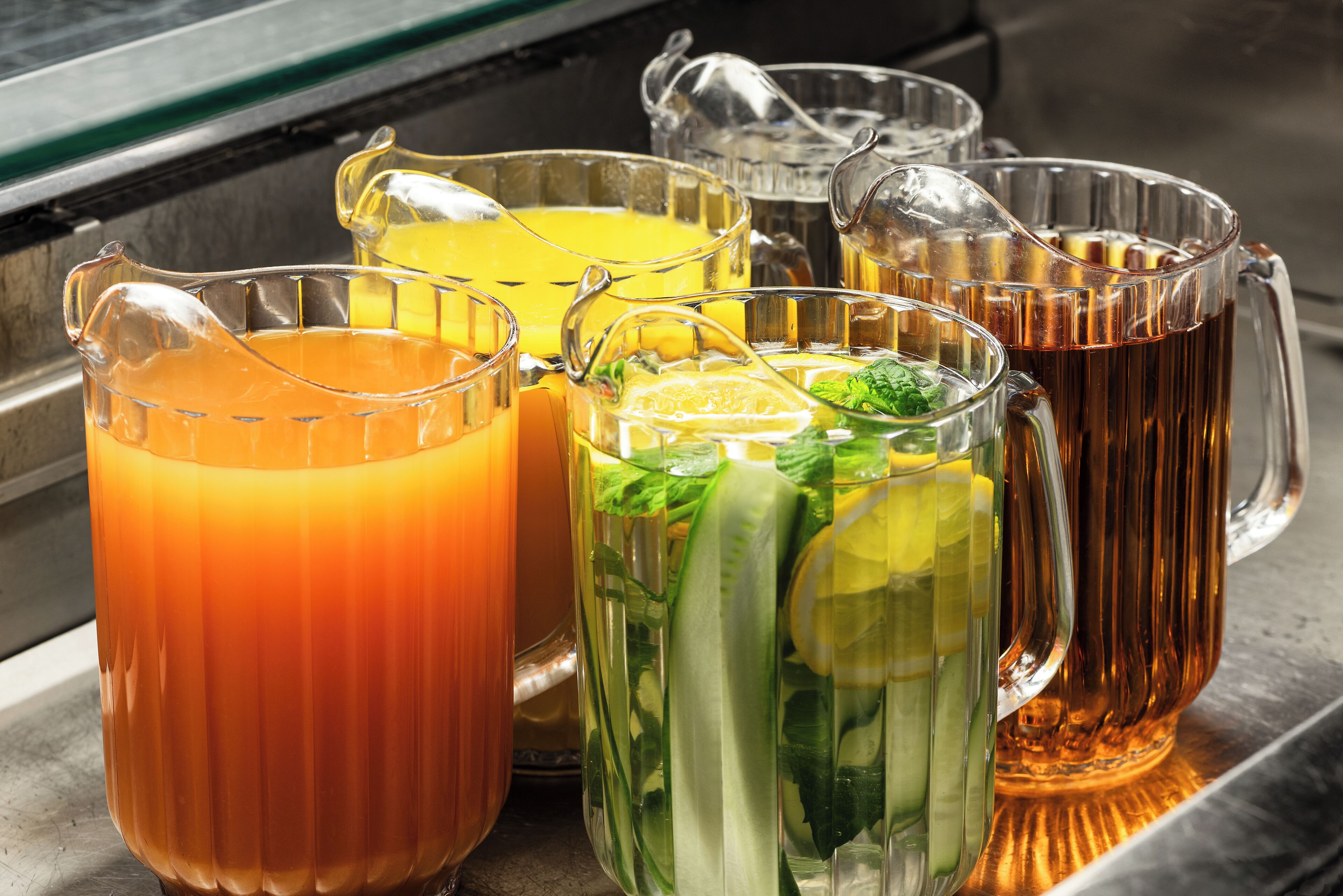 Diverse bevande in caraffe e tazze di vetro trasparente, alcune con ghiaccio e guarnizioni, su una superficie metallica.