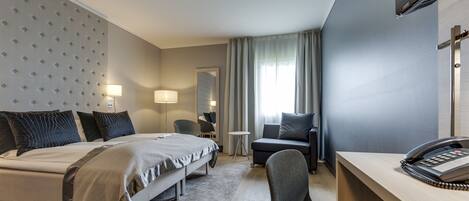 A hotel room with a bed, a desk, a chair, a small table, a lamp, a telephone, and a window with curtains.