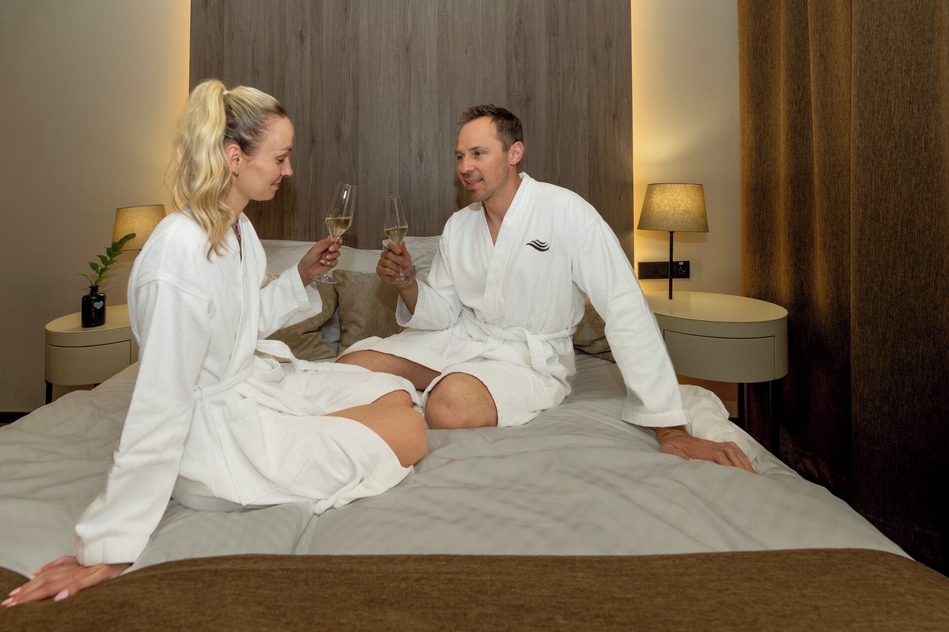 A couple in white robes sitting on a bed with a wooden headboard, holding wine glasses. There are bedside tables with lamps and a small plant on one side.