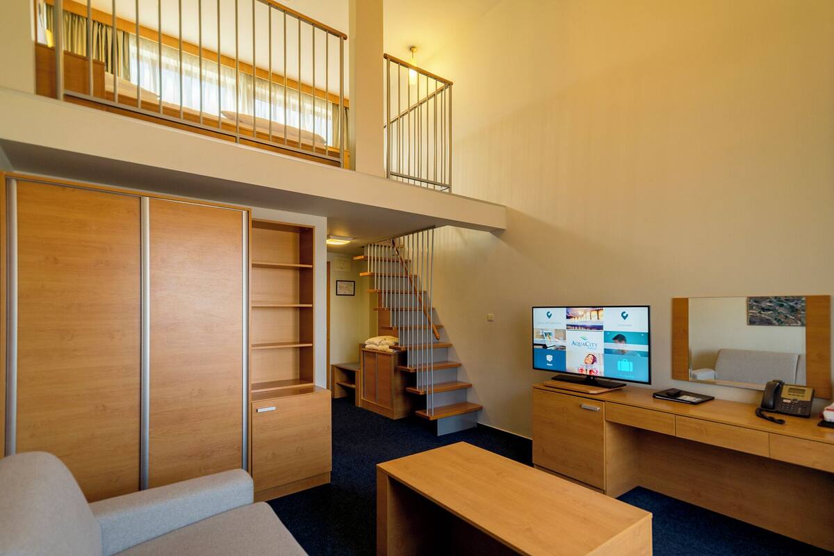 Ein kompaktes Hotelzimmer mit Schlafbereich auf Erhöhung, einem Schreibtisch inklusive Computer und einer kleinen Sitzecke.