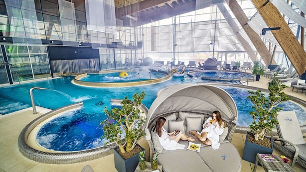 Une piscine intérieure dotée de plusieurs éléments aquatiques, notamment un jacuzzi et une piscine à vagues. On y trouve également des espaces de détente avec des sièges et des plantes en pot.