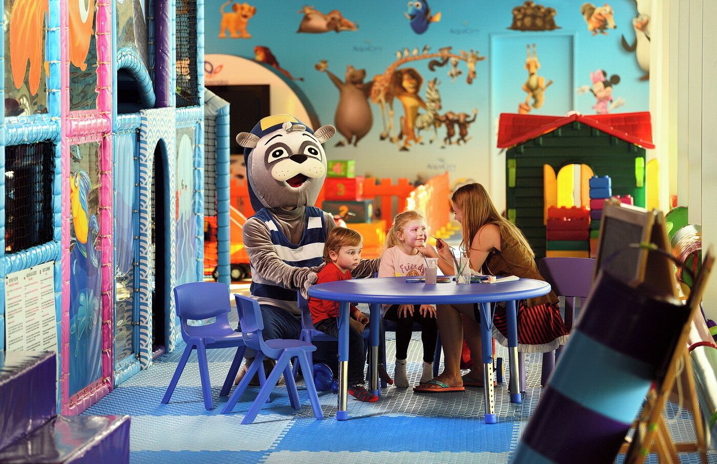 Un espace de jeux avec une mascotte, des enfants et des adultes assis autour d’une table.