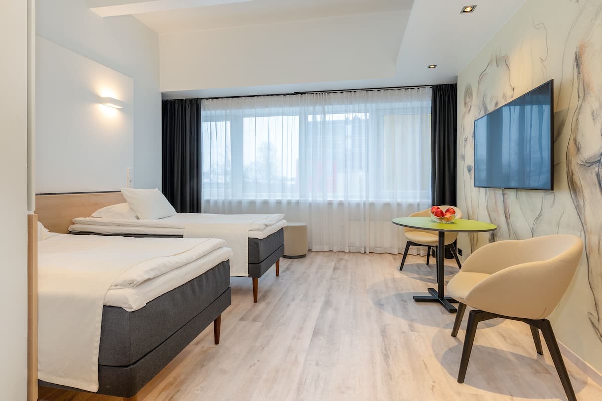 Ein Hotelzimmer mit zwei Betten, einem kleinen Tisch mit einer Obstschale, einem Stuhl und einem an der Wand befestigten Fernseher.