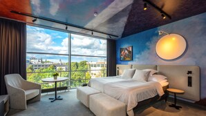 A modern hotel room with a large bed, a sofa, a small table with flowers, and a view of the city through large windows. - Art hotel Pallas by Tartuhotels (Tartu)