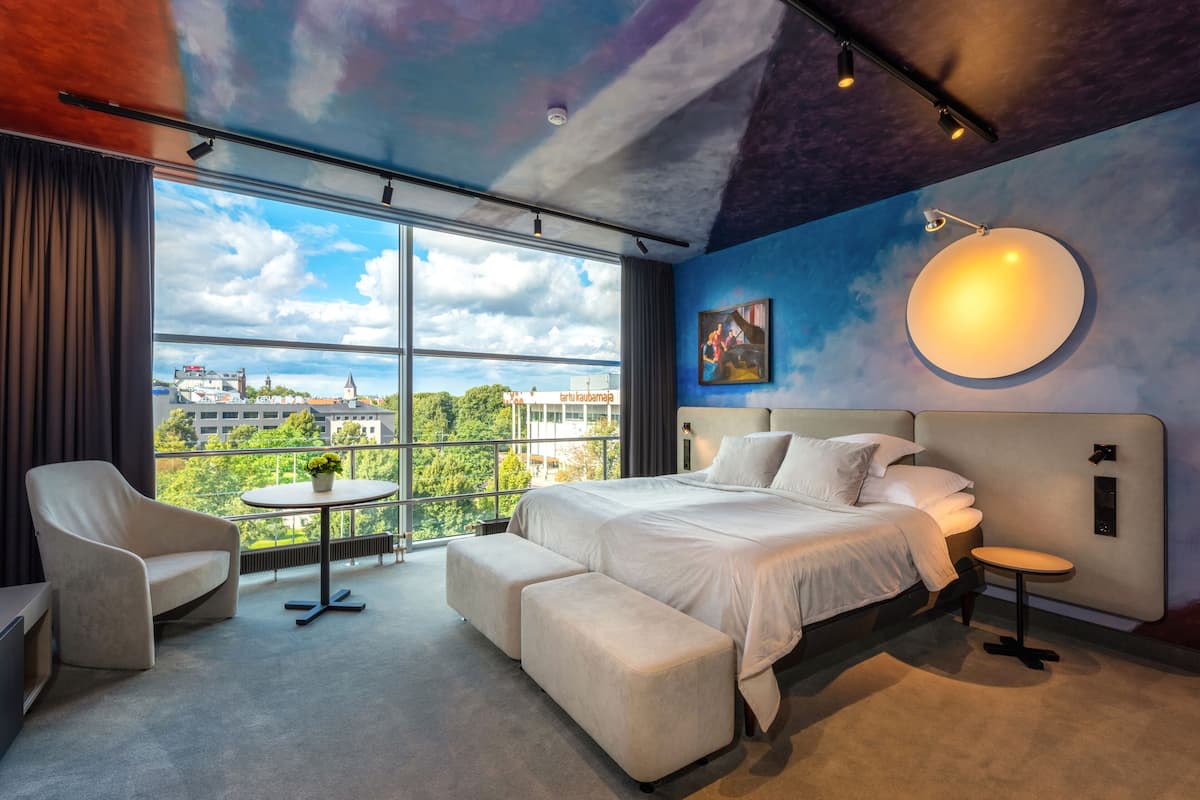 Ein modernes Hotelzimmer mit einem großen Bett, einer Sitzecke, einem kleinen Tisch mit Blumen und Blick auf die Stadt durch große Fenster.