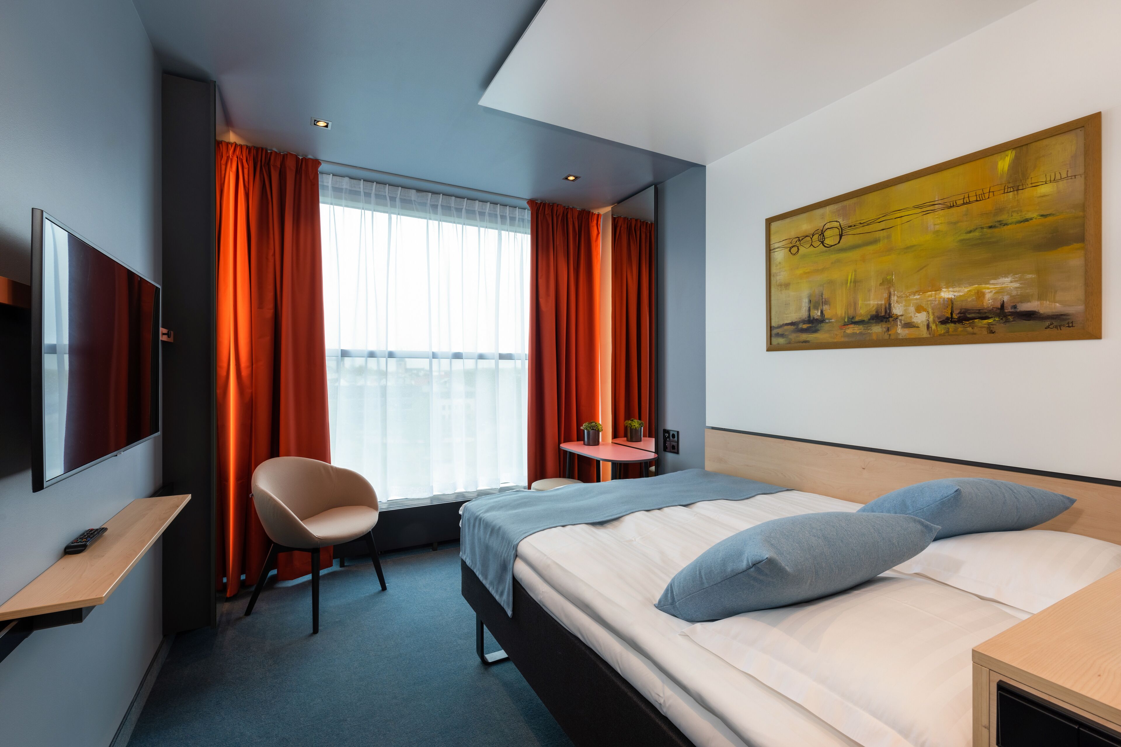 Ein Hotelzimmer mit einem Bett, einem Sessel, einem Schreibtisch, einem Fernseher und einem Bild an der Wand.