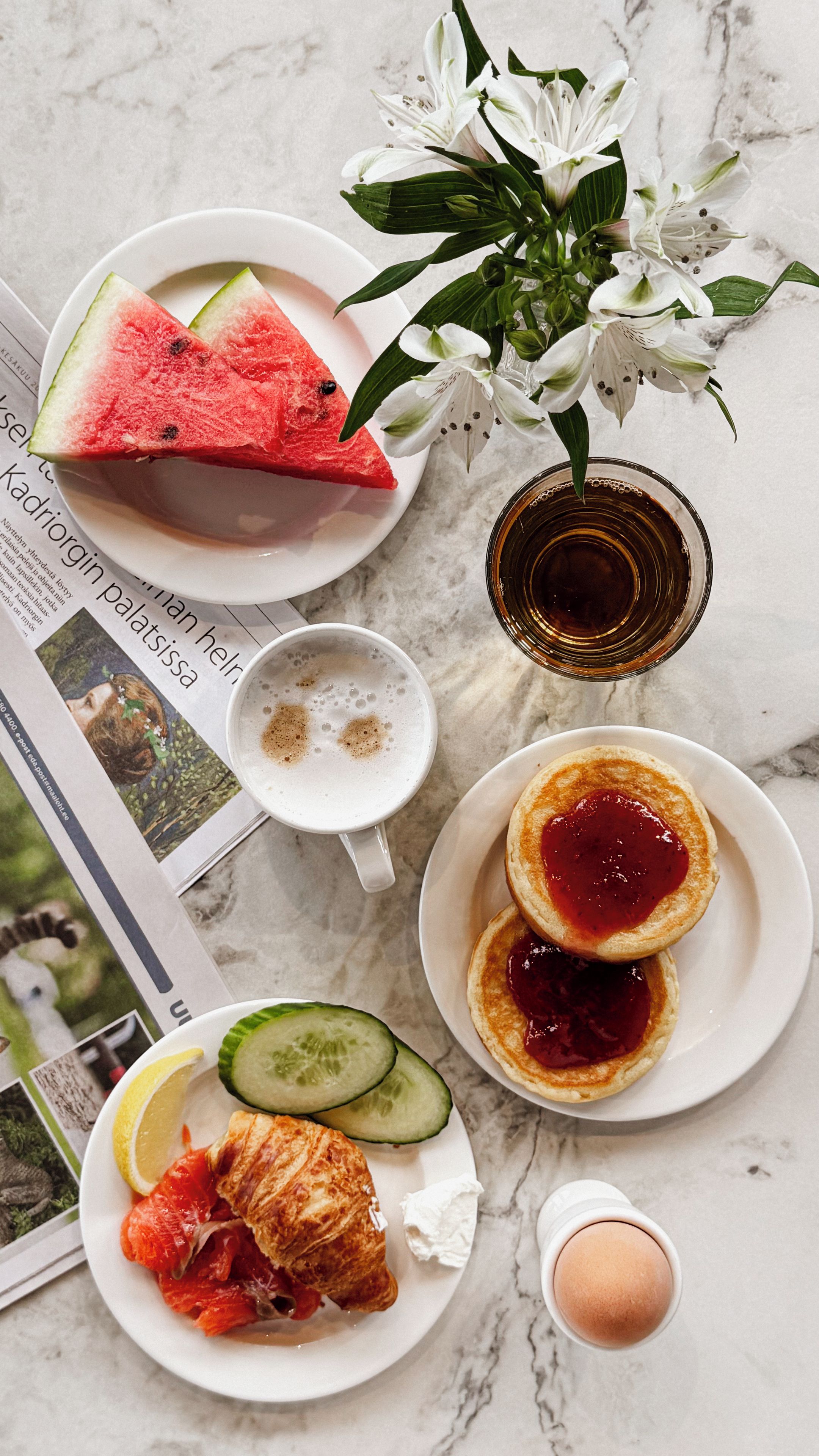 A breakfast setting with a plate of pancakes, a slice of watermelon, a cup of coffee, a glass of juice, a plate with a slice of lemon, a cucumber, and a plate with smoked salmon.