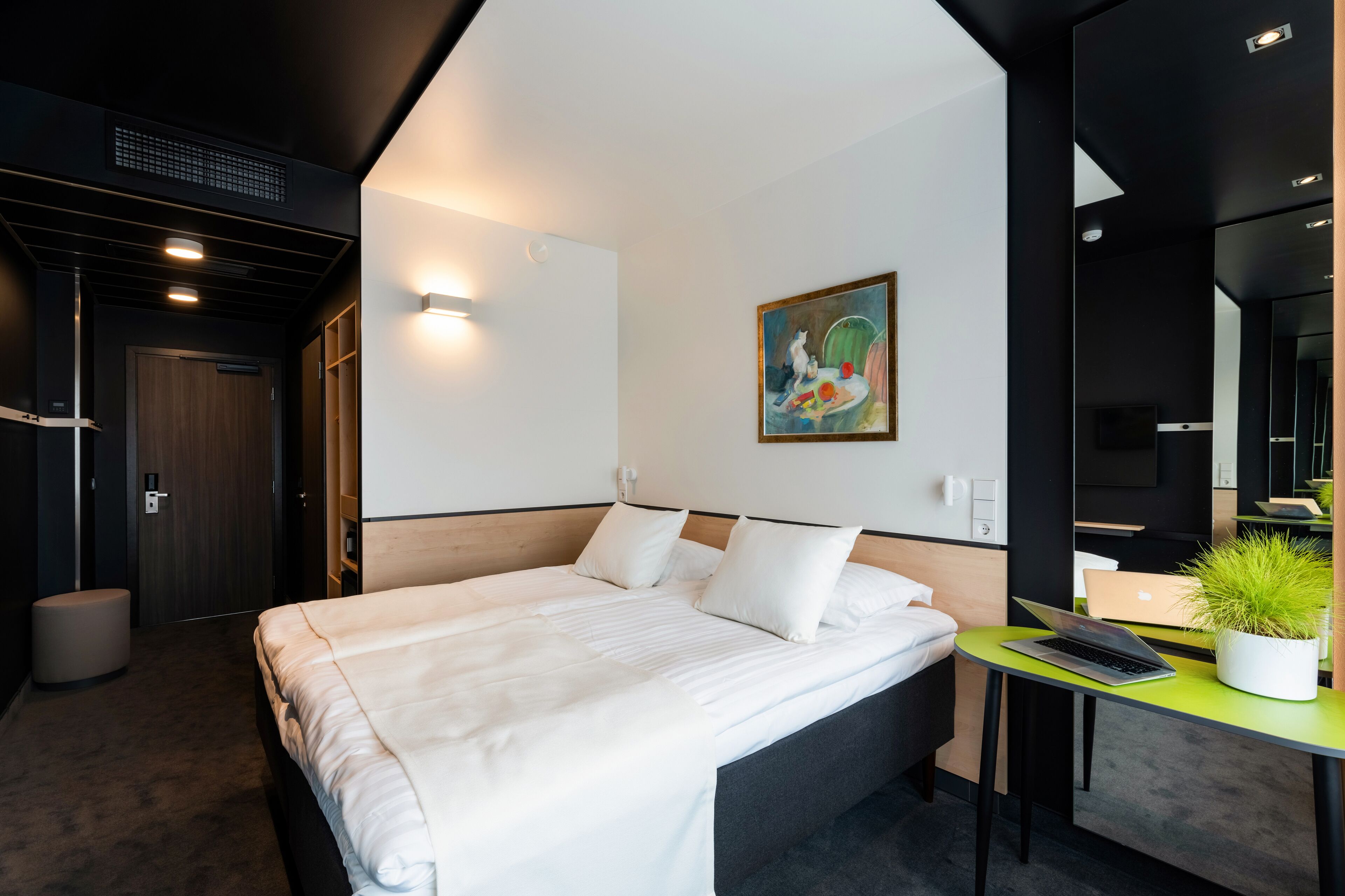 Et moderne hotelværelse med en stor seng, et skrivebord med en bærbar computer, en potteplante og et billede på væggen.