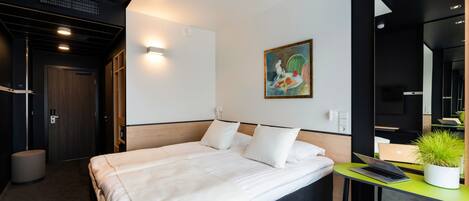 A modern hotel room with a large bed, a desk with a laptop, a potted plant, and a painting on the wall.