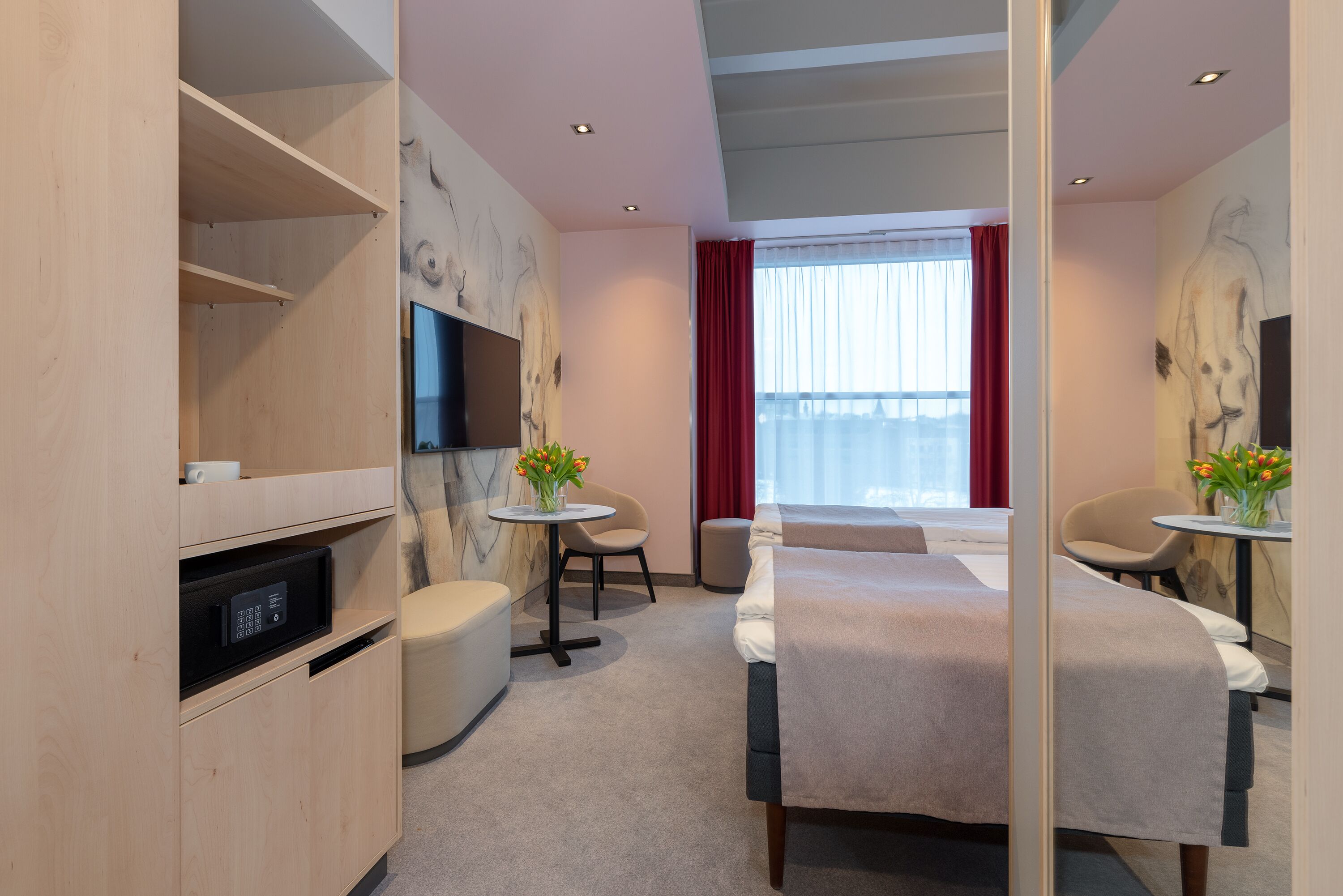 A hotel room with a bed, a desk, a chair, a TV, and a small table with flowers.
